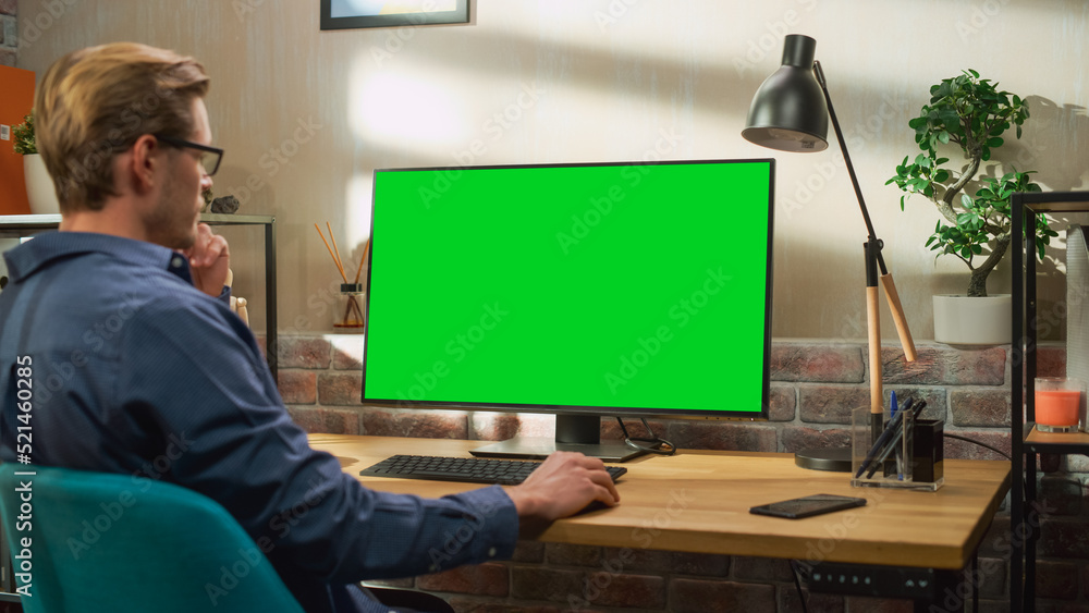 Poster Young Handsome Man Working from Home on Desktop Computer with Green Screen Mock Up Display. Creative Male Checking Social Media, Browsing Internet. Living Room in Bright Loft Apartment.