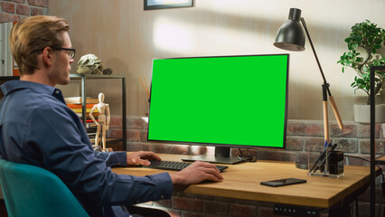 Young Handsome Man Working from Home on Desktop Computer with Green Screen Mock Up Display. Creative Male Checking Social Media, Browsing Internet. Living Room in Bright Loft Apartment.