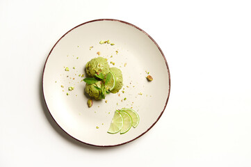 Flat lay. Food still life of three frozen green balls of pistachio and lime ice cream sorbet,sprinkled with pistachio nuts, melting on a plate, isolated on white background with copy space for text.