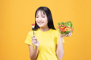 Portrait of happy asian woman eating tasty fresh vegetable salad holding plate bowl and fork on yellow background.
