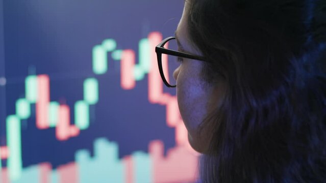 Growth of stock market. Portrait of focused woman trader wearing eyeglasses looking at a computer screen with trading charts. Back view. Concept of stock trading market, cryptocurrency and analyst.