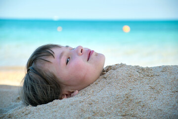 Young smiling child girl lying down covered with white sand on tropic beach on blue sky and ocean...