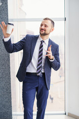 A young man with a neat little beard is dressed in a business suit with a white shirt and tie with a disposable paper cup of tea or coffee by the large window. Positive young businessman at the window