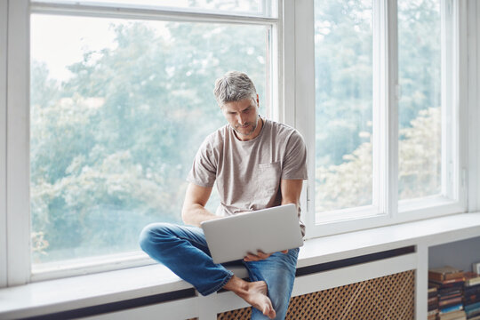 man using a laptop sitting on the windowsill in the living room.