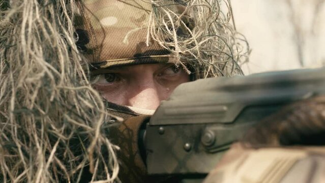 A close-up of a sniper wearing a camouflage suit or grass cloak, waiting and looking through a rifle scope. The sniper aims and shoots. Marksmanship shooting. Cinematic shot