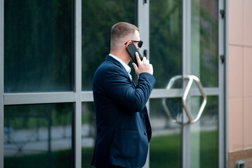 A business man in dark sunglasses with a phone in his hands stands outside the business building and talks on the phone. He is dressed in a light shirt and casual style jacket.