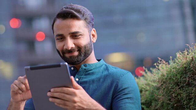 Smiling young adult indian business man professional, eastern businessman executive standing outdoors in urban city street reading ebook, holding using digital tablet online technology outside.