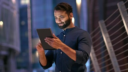 Smiling young indian business man professional, eastern businessman executive standing outdoors on street reading ebook holding using digital tablet online technology in night city with urbans lights. - Powered by Adobe
