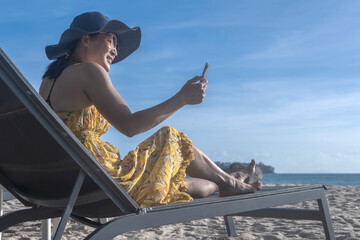 Asian female tourist sitting on beach chair using smartphone selfie herself in Phuket, Thailand, ...