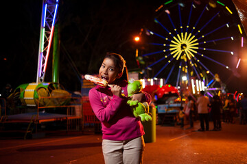 Smiling girl at fair eating corn in a cob with a Ferris wheel behind her. 