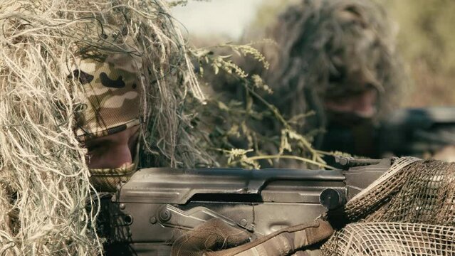 A close-up of a sniper wearing a camouflage suit or grass cloak, waiting and looking through a rifle scope. The sniper aims and shoots. Marksmanship shooting. Cinematic shot
