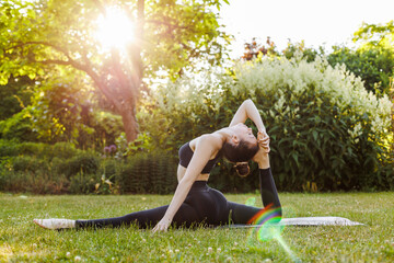 Young woman in black clothing practice yoga on mat in the summer park. Slim girl in sunset rays...