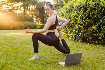 Young woman with laptop practice yoga in the summer park. Concept of outdoors online yoga classes, fitness learning or teaching, active and sporty lifestyle