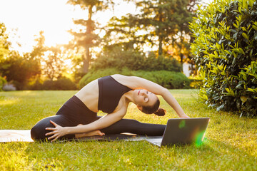 Young woman with laptop practice yoga in the summer park. Concept of outdoors online yoga classes, fitness learning or teaching, active and sporty lifestyle