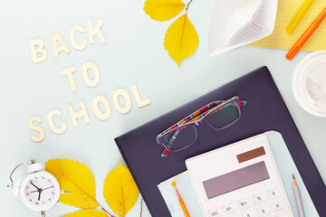 Back to school concept. School supplies on a tablet with glasses on a blue background with yellow, autumn leaves, alarm clock and wooden inscription Back to school. Flat lay