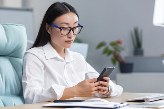 A Young Beautiful Asian Business Woman In Glasses Is Holding A Phone In Her Hands, Dialing A Message. Sitting At A Desk In A Modern Office.