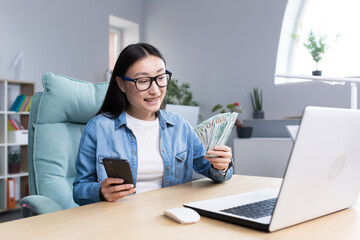 Fototapeta premium Business training about money. A young Asian business woman conducts a training session on the topic of money. He is holding a phone and a wad of bills. Sits at his desk, talks, looks into the camera.
