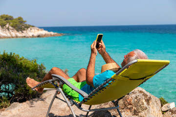 Mature man on holiday relax in the sun on the deck chair holding his phone chatting with friends.Vacation and technology.