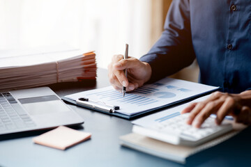 Businessman is using a calculator to calculate company financial figures from earnings papers, a businessman sitting in his office where the company financial chart is placed. Banner with copy space.