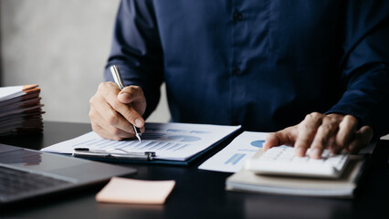 Businessman is using a calculator to calculate company financial figures from earnings papers, a businessman sitting in his office where the company financial chart is placed. Banner with copy space.