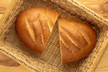 Two halves of a fragrant fresh long loaf in a straw basket on a wooden table, close-up, top view.