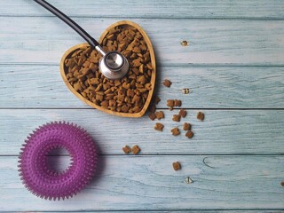 Dry pet food in heart shaped bowl with a stethoscope and a toy on a wooden table. Concept care for your pet.