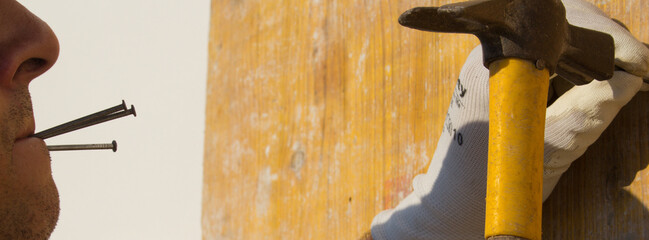 Image of a carpenter's mouth holding nails between his lips while nailing wooden boards with a...