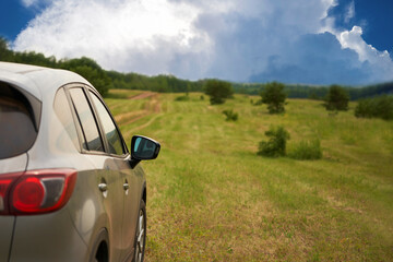 A fragment of a car moving along a field road through a meadow with mown grass. There is a hilly forested terrain on the horizon. Copy space.