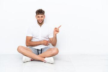 Young blonde man sitting on the floor isolated on white background pointing finger to the side