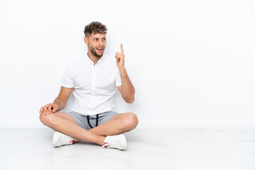 Young blonde man sitting on the floor isolated on white background intending to realizes the solution while lifting a finger up