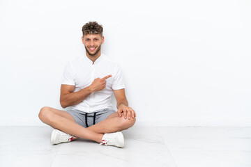 Young blonde man sitting on the floor isolated on white background pointing to the side to present a product