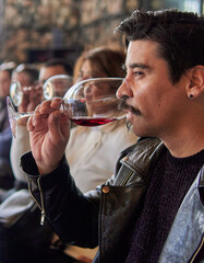 latin man in profile at a wine tasting at a winery in mendoza argentina. Other people blurred on background. Brunette man in leather jacket holding and savoring a glass of red wine.