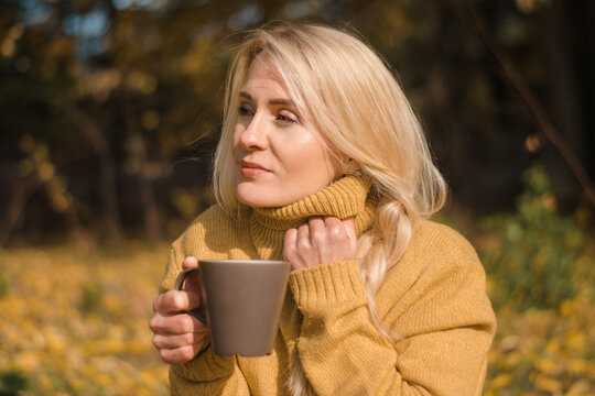 Attractive blonde woman in a yellow sweater is drinking tea at a picnic in an autumn park.Autumn concept.Beauty in nature.Slow living.