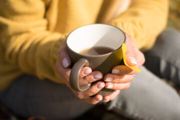Yellow leaf and mug with hot tea in female hands, close-up.The woman is dressed in a cozy yellow...