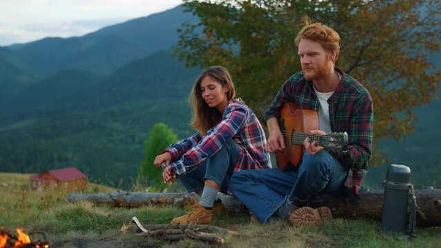 Positive couple date outdoors in mountains. Two lovers play guitar by campfire.