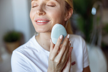 Portrait of young beautiful blonde Caucasian female massaging face skin with massager.