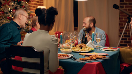 Multiethnic people gathered in living room to celebrate winter holiday while giving each other gifts. Festive family members sitting at Christmas dinner table while exchanging presents at home.