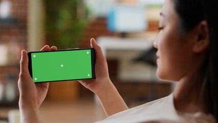 Smiling woman having smartphone with green screen chroma key background. Happy asian person with mobile phone having mockup isolated display while sitting on sofa at home.