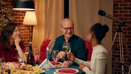 Festive smiling senior man clinking glasses with young woman at Christmas dinner table. Happy elderly person talking with family member while celebrating traditional winter holiday at home.
