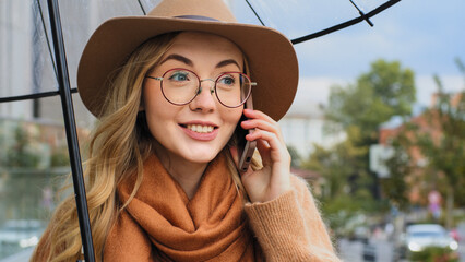 Attractive young woman with umbrella answers call on the street during rain, smart girl...