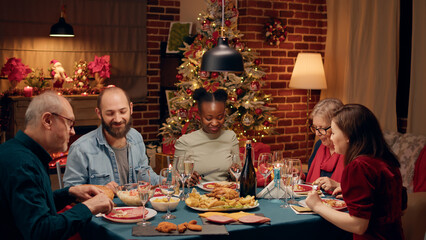 Festive multiethnic family enjoying traditional home cooked food while sitting at Christmas dinner table. Happy people at home celebrating winter holiday together while being positive and peaceful.