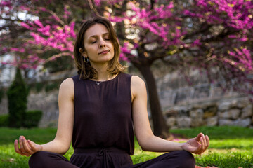 Brunette Woman with Earrings Breathing Deeply with Closed Eyes in Meditation Posture in a Public Park with Green Grass and a Purple Leaved Tree Behind by Spring