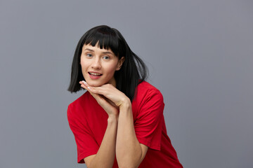 a sweet, modest, attractive woman stands on a gray background in a red T-shirt and smiling pleasantly at the camera keeps her fingers folded in a relaxed pose