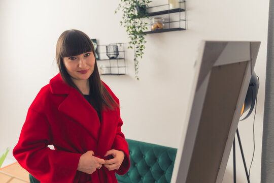 Charming Young Brunette Woman Putting On A Red Coat In Front Of The Mirror In Her Apartment. High-quality Photo