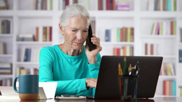 Senior elderly woman working on laptop computer while talking on smartphone.