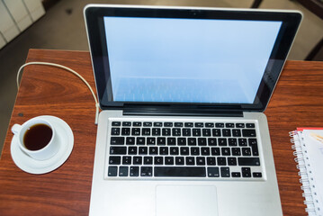 open laptop on a wooden table, coffee cup and blank notebook next to it
