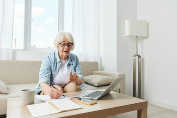 Portrait of an elderly business woman taking notes in a notebook sitting on the couch at home with a laptop undergoing online training