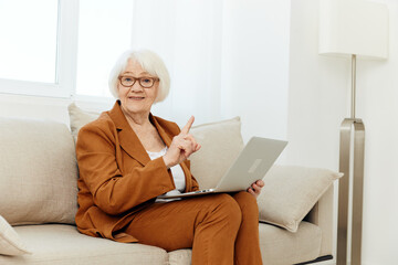 attentive elderly woman in a classic suit in a bright room on the sofa and working on a laptop remotely