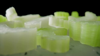 Sliced slices of fresh juicy celery stalk on a gray plastic board. Macro shot of chopped green celery pieces on black isolated studio background. Ingredient for salad. Slow motion ready 59.94fps.