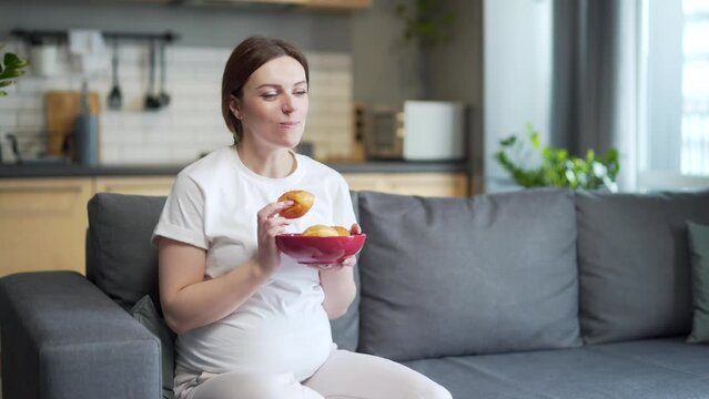 Hungry pregnant woman with belly eats sweets unhealthy and junk food. Female with an appetite hold plate and eating cookies donut. Sitting at home on the sofa. Indoor. Sweet cravings during pregnancy